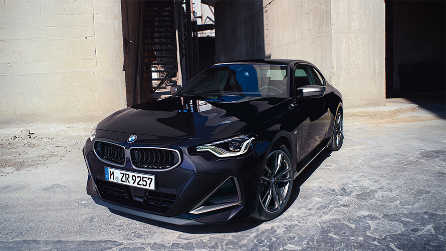 BMW M240i xDrive Coupé G42 2021 Vista de tres cuartos del frontal, parado BMW M240i xDrive Coupé G42 2021 Black Sapphire metalizado Vista de tres cuartos del frontal, parado delante de un almacén