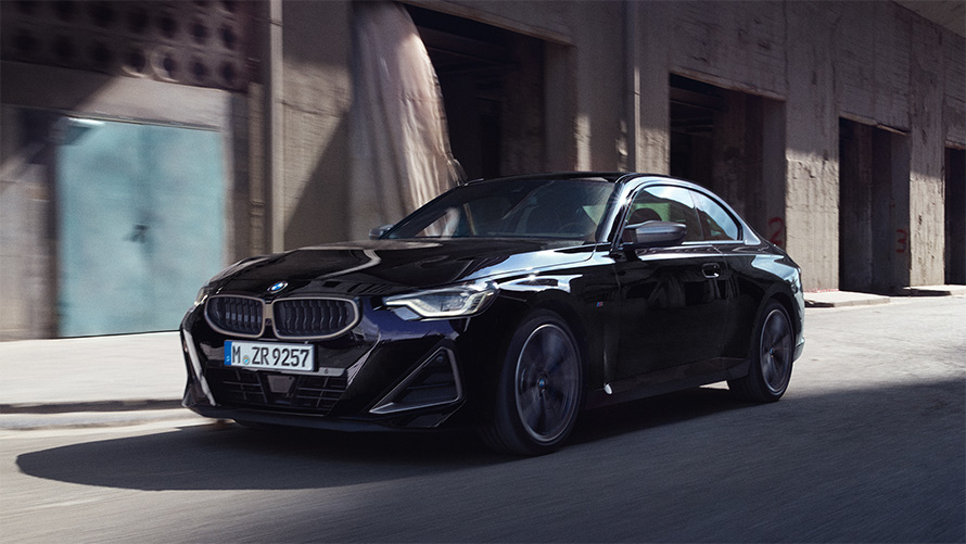 BMW M240i xDrive Coupé G42 2021 Vista de tres cuartos del lateral, en marcha BMW M240i xDrive Coupé G42 2021 Black Sapphire metalizado Vista de tres cuartos del lateral, en marcha delante de un almacén