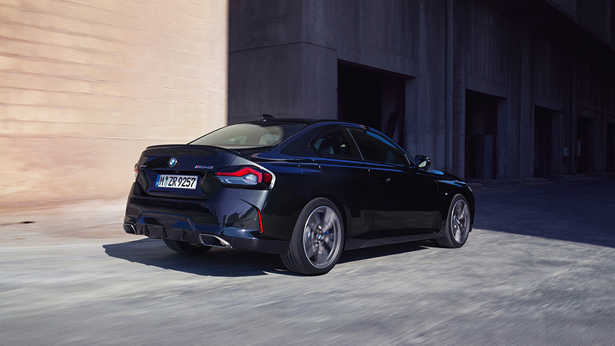 BMW M240i xDrive Coupé G42 2021 Vista de tres cuartos de la zaga, parado BMW M240i xDrive Coupé G42 2021 Black Sapphire metalizado Vista de tres cuartos de la zaga, parado delante de un almacén