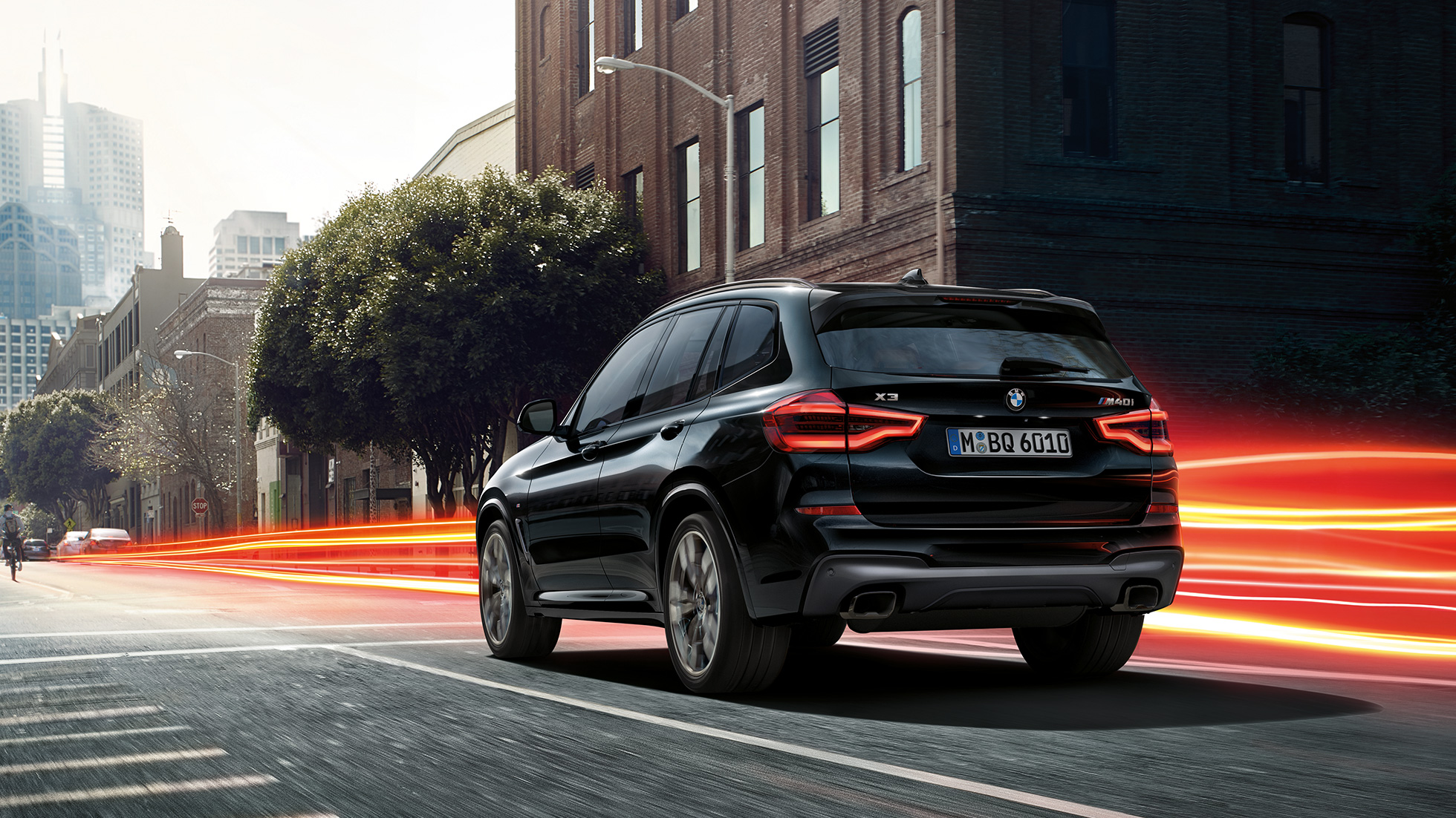 BMW X3 M40i, vista trasera de tres cuartos. BMW X3 M40i en Black Sapphire metálico, en movimiento, exterior, vista trasera de tres cuartos frente a un fondo urbano con franjas de luz.